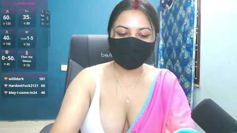 Video of a South Asian woman with medium skin tone, dark hair, and a black mask, wearing a pink and white sari. Background shows a blue wall and a weather forecast screen.