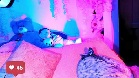 Video of a child lying on a bed covered with stuffed animals and pillows, bathed in vibrant blue and pink lighting.