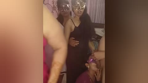 Video of three women in a dimly lit room, one wearing a pink dress and a tiara, another with a black dress and silver mask, and a third with a purple dress, all smiling.