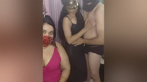 A video of three people in a dimly lit room: a woman in a red mask, a woman in a black dress, and a man topless, wearing black shorts.