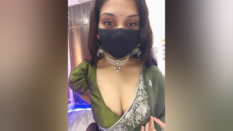 Video of a South Asian woman with medium-brown skin, wearing a green saree with white floral embroidery, black face mask, and silver jewelry, indoors.