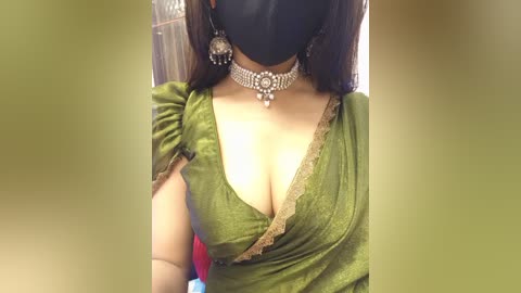 Video of a woman in a green, low-cut sari with lace trim, wearing a black mask, silver choker, and dangling earrings. Background features a blurred, beige curtain.