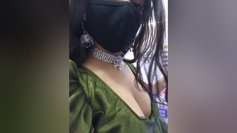 Video of a person with long, dark hair, wearing a black mask, a green glittery top, and a silver choker with a pendant, indoors with blurred background.