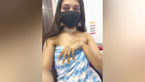 Video of a South Asian woman with long black hair, wearing a black face mask, blue tie-dye saree, and silver jewelry, standing indoors with a blurred background.