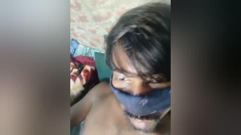 A close-up video shows a man with short dark hair, wearing a black blindfold and possibly shirtless, lying on a bed with a red and yellow patterned blanket.