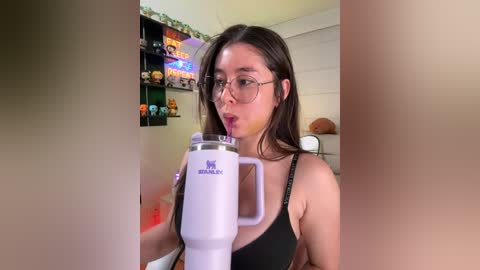 Video of a young Asian woman with glasses, wearing a black tank top, drinking from a white blender, surrounded by colorful toy figurines and shelves in a cluttered room.