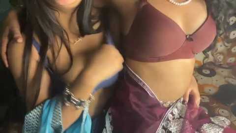 Video of two women in lingerie, one in a blue bra and the other in a burgundy bra, both with medium-sized breasts, standing closely in a dimly lit room with a floral-patterned background.