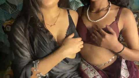 Video of two women in traditional Indian attire, with one in a maroon blouse and the other in a black saree, both wearing pearl necklaces.