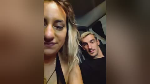 Video of a young woman with long, wavy blonde hair and a light complexion, wearing a black top, and a man with short, light brown hair in the background, both smiling.