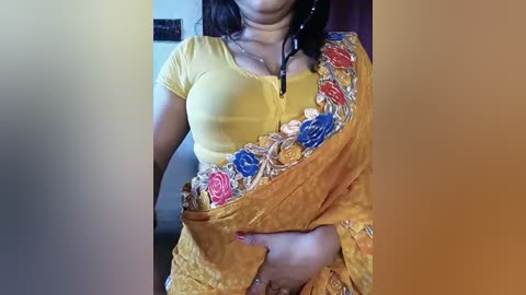 Video of a woman in a yellow blouse, adorned with a floral-patterned sari, featuring intricate embroidery in red, blue, and white. She wears a black necklace. Background shows a blurred indoor setting with muted colors.