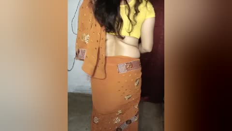Video of a woman with long dark hair, wearing a yellow crop top and a brown sari adorned with floral patterns, standing indoors near a maroon curtain.