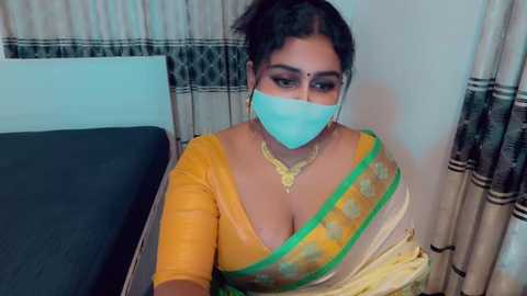 Video of a South Asian woman with medium brown skin, wearing a yellow saree, green border, and a light blue mask, seated on a bed with beige curtains and a dark headboard.