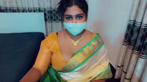 Video of a South Asian woman with dark hair, wearing a yellow and green saree, blue mask, gold necklace, and sitting on a bed with beige curtains in the background.
