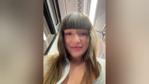 Video of a young woman with long, straight, light brown hair styled in a blunt bob cut. She has a light skin tone and is wearing a light grey shirt. The background features a subway train interior with metallic surfaces and a patterned wall.