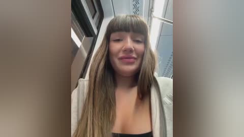 Video of a smiling woman with long, straight brown hair and blunt bangs, wearing a white cardigan over a black top, seated in a train compartment with metal railings and white walls in the background.