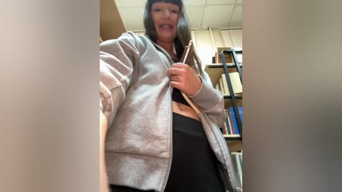 A video of a middle-aged woman with straight gray hair, wearing a gray zip-up hoodie and black leggings, standing in a library with shelves of books in the background.