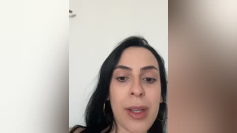 Video of a young woman with medium skin tone, long black hair, and full eyebrows, wearing minimal makeup, standing against a white and beige wall, smiling slightly.