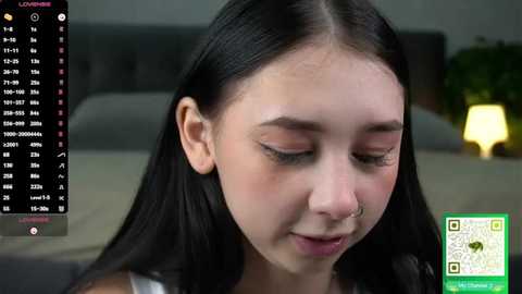 A video of a young woman with long black hair, fair skin, and a nose ring, smiling gently. Background shows a blurry bed and green plant. Overlay shows social media app screen.