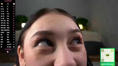 Video of a young woman with fair skin, brown eyes, and long brown hair, looking upward. Background shows a green plant and a calendar with dates. QR code in bottom right.