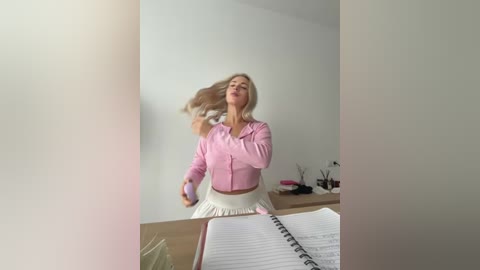 Video of a blonde woman with long hair, wearing a pink button-up shirt and white skirt, dancing joyfully in a minimalist office setting.