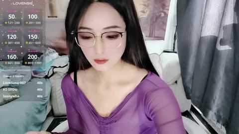 Video of an East Asian woman with long black hair, wearing glasses and a sheer purple top, sitting indoors. Digital screen overlay displays social media metrics. Background includes a bed with white sheets and a grey curtain.