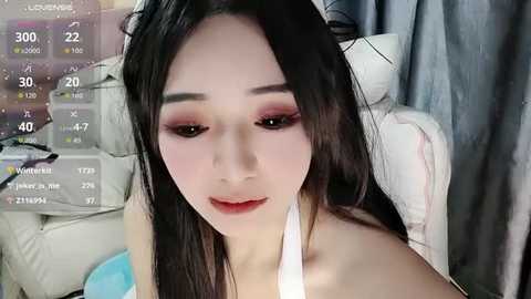 A video of a young East Asian woman with long black hair, wearing makeup, in a white top, sitting in a modern room with gray curtains and digital camera overlay.
