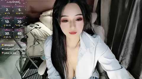 Video of a young East Asian woman with long black hair, fair skin, and prominent makeup, wearing a low-cut white jacket, seated on a bed with a digital clock displaying 40\u00b0C.