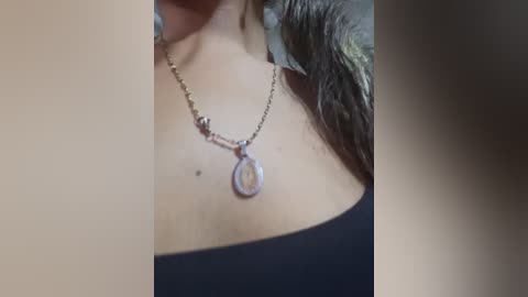 A video of a woman's neck and upper chest showing a silver necklace with a circular pendant. She is wearing a black off-the-shoulder top. Background is blurry.