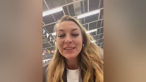 Video of a smiling, light-skinned woman with long, wavy blonde hair and light makeup, wearing a white shirt, in an indoor setting with fluorescent lighting and white ceiling panels.