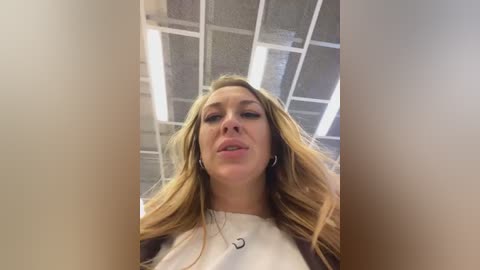 A video of a Caucasian woman with long blonde hair, wearing a white top, standing in an office with a ceiling of fluorescent lights and white tiles.