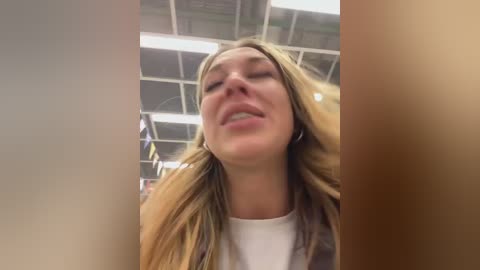 Video of a young woman with long, blonde hair, wearing a white top, smiling widely. She is indoors with a ceiling light and blurred background.