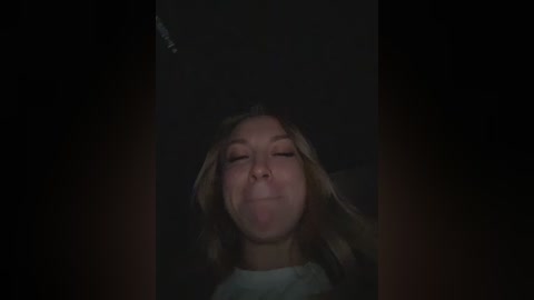 A dimly lit video captures a Caucasian woman with light brown hair, closed eyes, and a relaxed expression, her hands gently resting on her head. The background is dark, suggesting a nighttime setting.