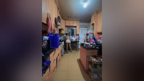 Video of a cluttered, dimly lit kitchen with beige cabinets, a man wearing a white tank top and shorts, and various cooking utensils scattered around.