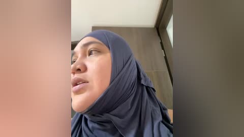 Video of a light-skinned, young woman with a round face and short, neatly styled hair, wearing a dark blue hijab. She is gazing upward with a neutral expression. Background features modern, minimalist interior design with wooden furniture.