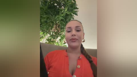 Video of a smiling, light-skinned woman with braided hair, wearing a red blouse, sitting indoors next to a green plant.