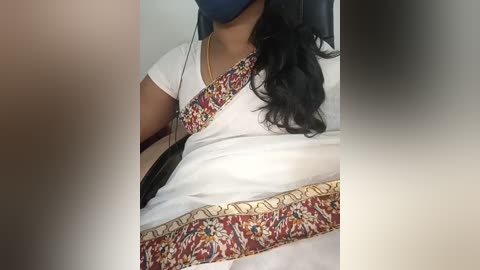 A video showing a person wearing a white short-sleeved shirt with a colorful patterned sash, a blue mask, and a black bag slung over the shoulder, seated in a car.