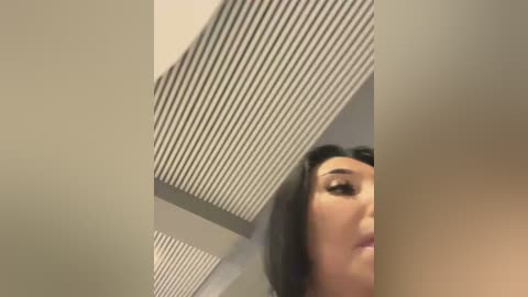 A close-up video shows a young woman with long black hair, partially obscured by a vertical white pillar, under a grid-patterned ceiling. Her face is partially visible, highlighting her light skin and delicate features.