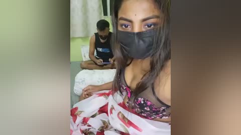 Video of a South Asian woman with medium skin tone, wearing a floral-patterned dress, black face mask, and floral bra, sitting on a bed, while a man in a black tank top and white mask sits on the floor, reading a book in a dimly lit room.