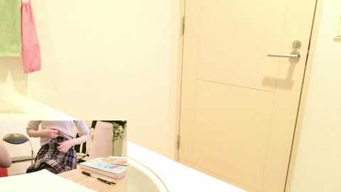 Video of a minimalist bathroom with a beige door, a white sink, and a towel rack holding a pink towel. A small, partially visible mirror image of a person in a white shirt and plaid shorts is reflected on the sink.