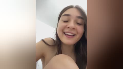 Video of a young Latina woman with long dark hair, medium skin tone, smiling, topless, standing in a dimly lit room.
