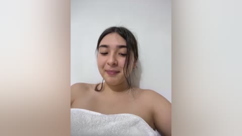 Video of a young Latina woman with medium-brown skin, dark hair, and a slight smile, wrapped in a white towel, standing against a plain white wall, viewed from the side.