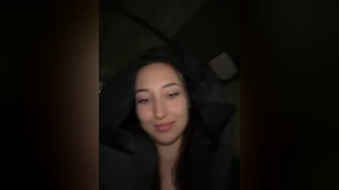 Video of a woman with light skin, straight dark hair, wearing a black jacket, smiling softly, in dimly lit, possibly indoor setting.