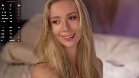 Video of a blonde woman with long hair, fair skin, and blue eyes, smiling in a bedroom with a bed and soft lighting.