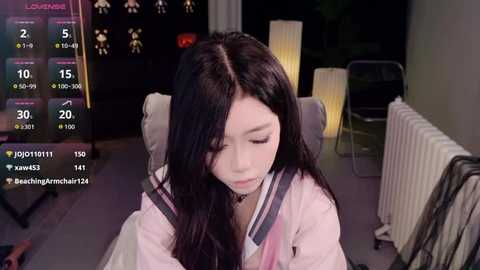 Video of a young Asian woman with long black hair, wearing a pink robe, sitting in a dimly lit room with a radiator, modern furniture, and digital displays showing time and temperature.