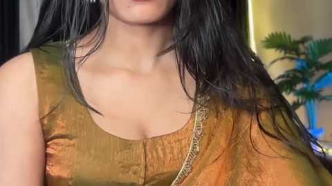 Video of a woman with long, straight black hair, wearing a mustard-yellow blouse with intricate embroidery, indoors with a blurred background.