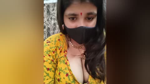 Media: Video of a South Asian woman with dark skin, wearing a yellow floral blouse, black face mask, and red dot on forehead, indoors.