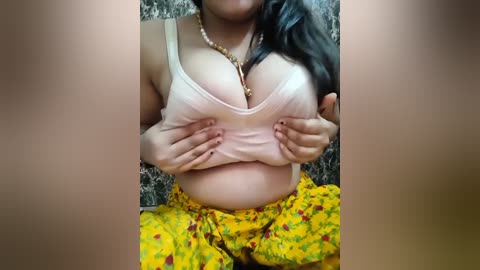 Media: Video of a light-skinned woman with long dark hair, wearing a white crop top lifted to expose ample cleavage, and a yellow floral skirt. She is indoors, seated on a textured surface.