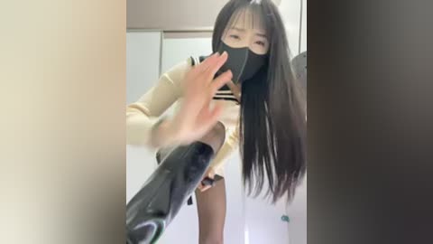 Video of an Asian woman with long black hair, wearing a beige sweater and black face mask, holding a black high-heeled shoe.