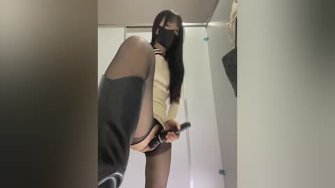 A video of an Asian woman with long black hair, wearing a beige sweater, black skirt, and knee-high socks, holding a black object, in a dimly lit bathroom.