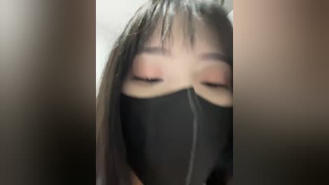 A close-up video of an Asian woman with long black hair and a black face mask, eyes closed, wearing pink eyeshadow and blush, set against a blurred background.